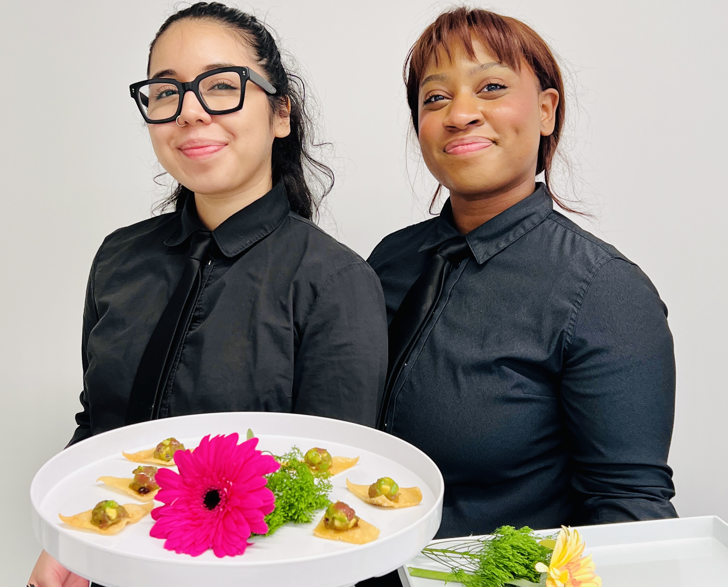 Two smiling Sweet Basil Catering staff members in formal black uniforms holding trays of gourmet hors d'oeuvres, beautifully garnished with fresh greens and a pink flower, during a corporate event.