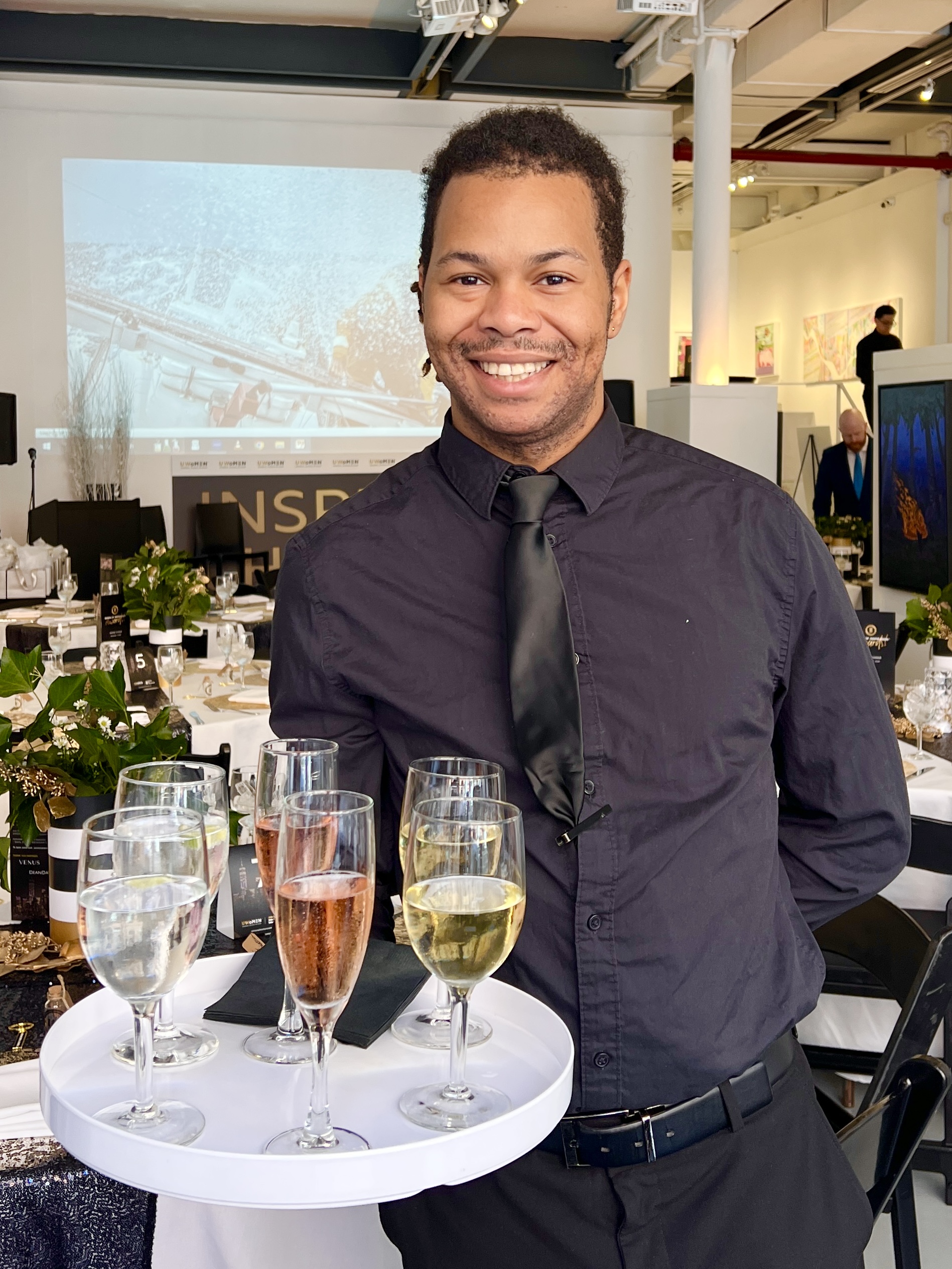 A smiling Sweet Basil Catering staff member holding a tray of wine and champagne glasses, set against an elegant corporate event space.