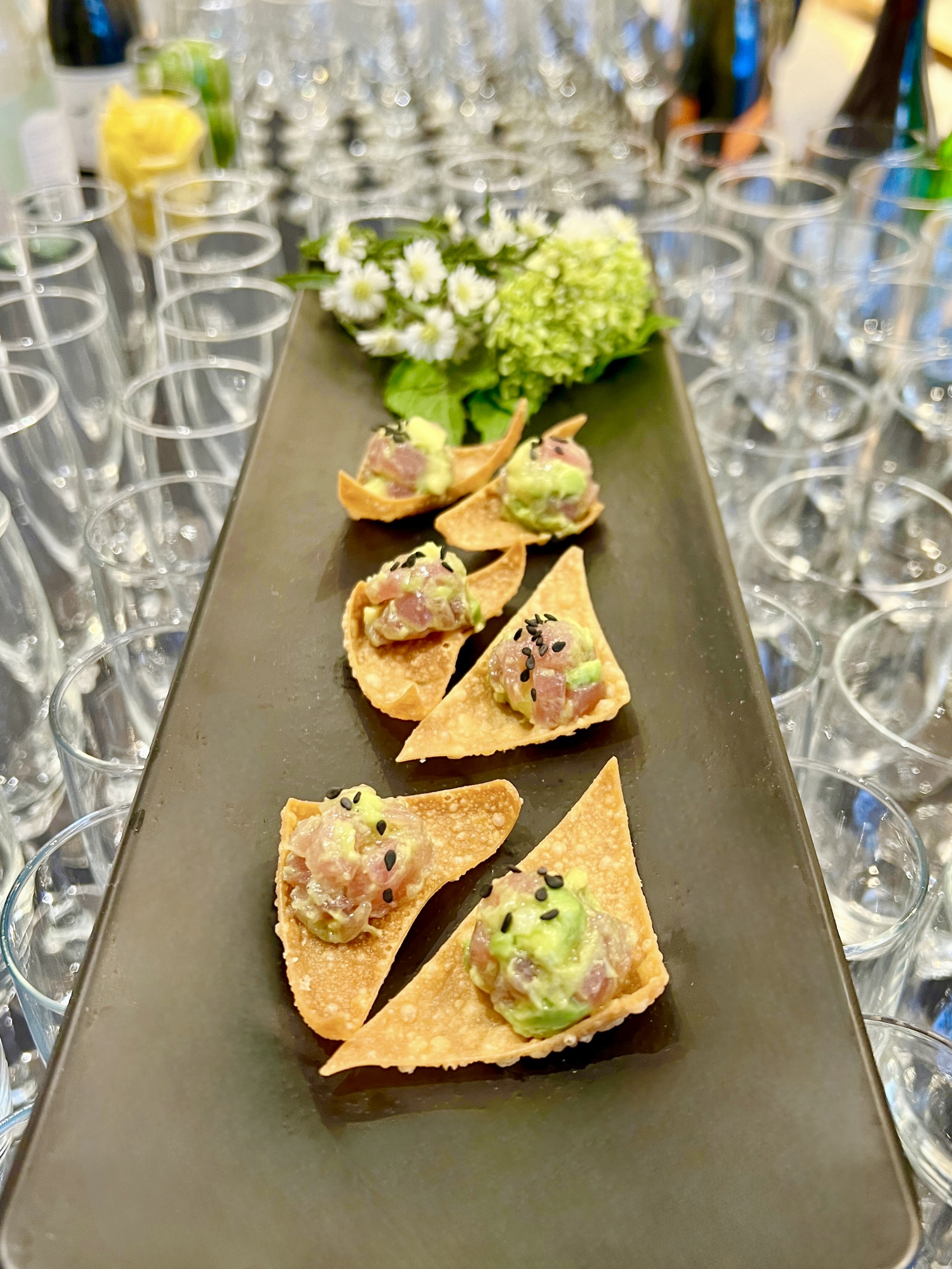 A close-up of Sweet Basil Catering's tuna tartare bites on crispy wonton chips, garnished with avocado and sesame seeds, displayed on a black tray with fresh flowers.