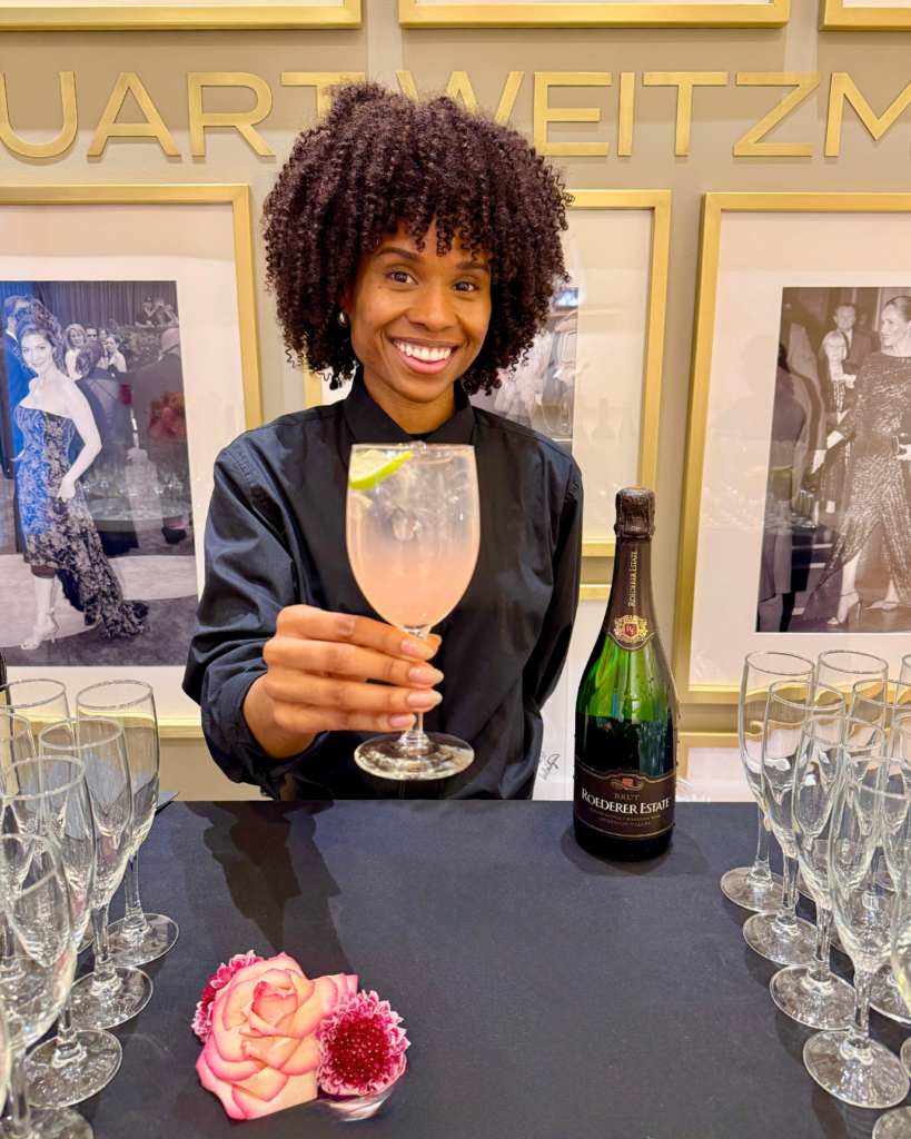 Bespoke Bar Catering NYC - A smiling bartender in a black uniform holds out a cocktail garnished with a lime wedge at an elegant event. The bar setup includes champagne glasses, a bottle of Roederer Estate Brut, and decorative pink and red flowers on a black tablecloth. The background features framed black-and-white fashion photographs and gold-lettered branding on the wall.