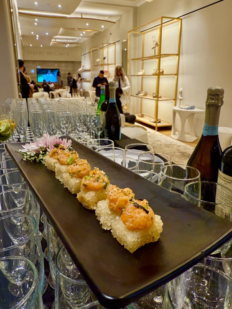 A beautifully presented plate of crispy rice topped with spicy tuna, garnished with sesame seeds and chives, served at the Stuart Weitzman + Power Haus Women event in New York City. The dish is placed on a sleek black platter over a champagne station, with the elegant event space in the background.