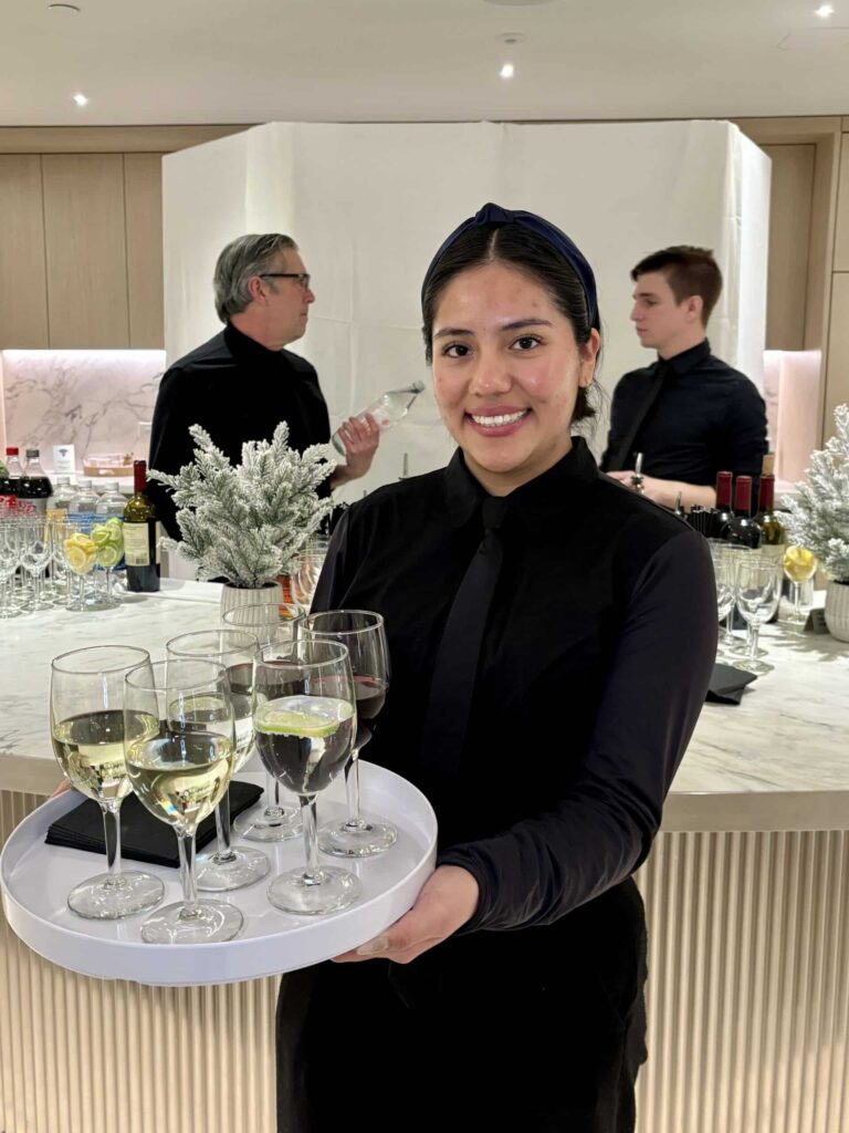 Smiling server holding a tray of wine glasses in front of a modern event bar, with two bartenders preparing drinks in the background.