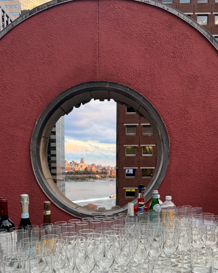 Outdoor bar setup with glassware and liquor bottles, overlooking the New York City skyline through a circular red brick window.