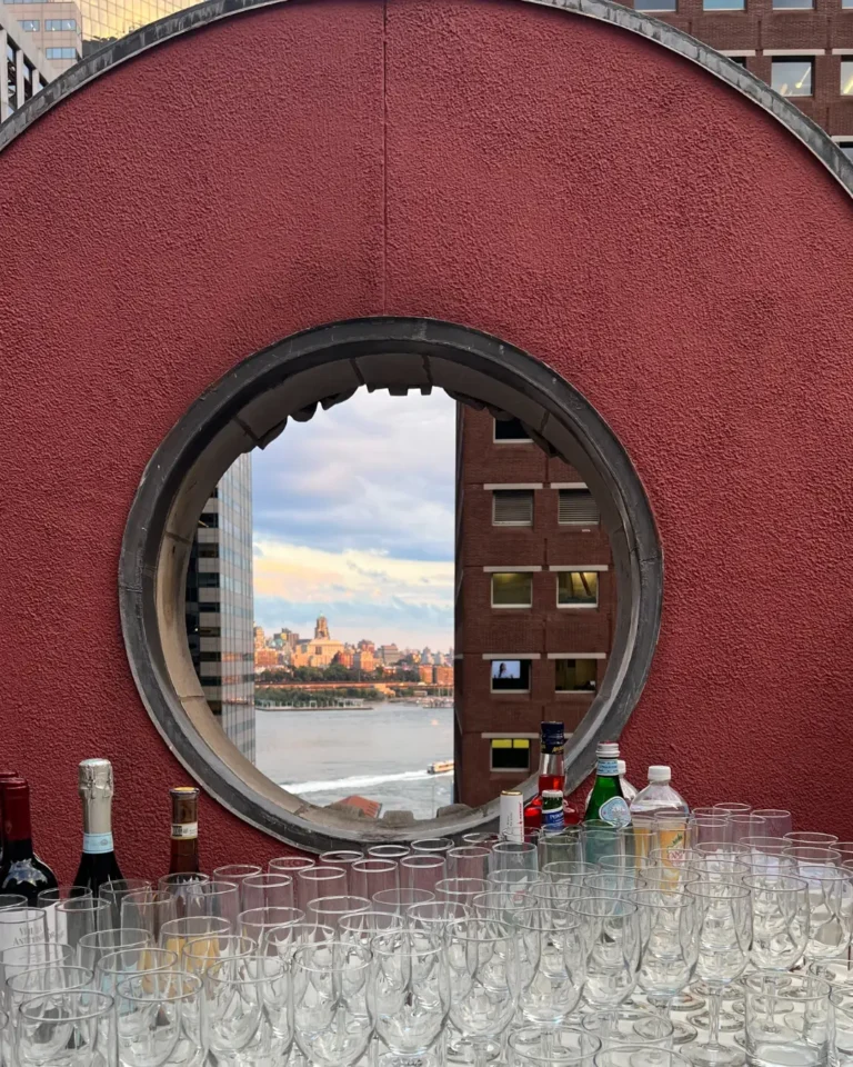 Outdoor bar setup with glassware and liquor bottles, overlooking the New York City skyline through a circular red brick window.