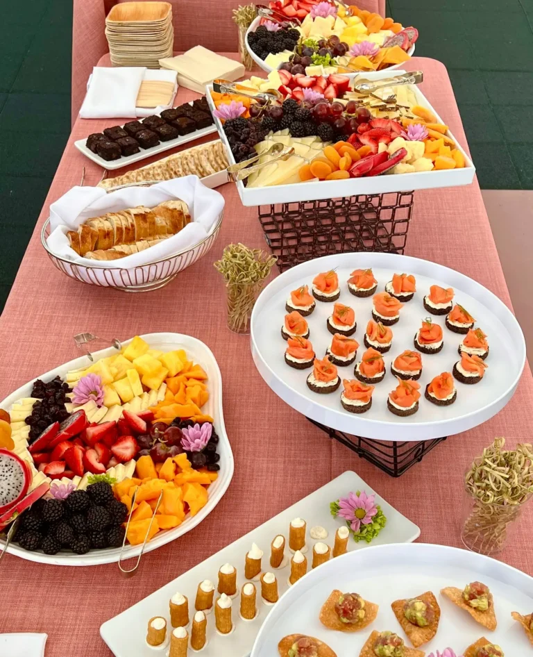 Colorful grazing table setup with assorted fruits, cheeses, smoked salmon bites, and mini desserts on a pink tablecloth at a rooftop event.