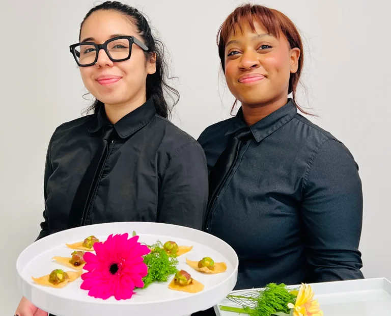 Two Sweet Basil Catering staff smiling while holding appetizer trays at a corporate event in NYC
